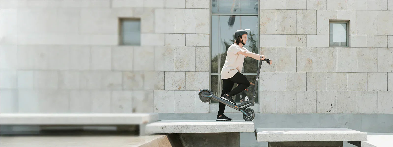 Eine Person, die einen Helm trägt, fährt mit einem Elektroroller eine Stufe in einem städtischen Außenbereich mit Steinmauern und Fenstern hinauf.