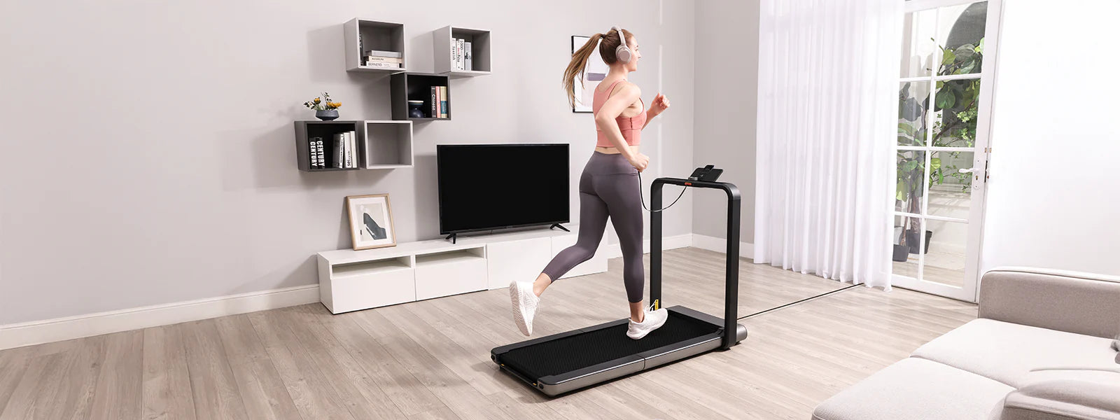 Eine Frau mit Kopfhörern läuft auf einem Laufband in einem modernen Wohnzimmer mit einem Fernseher, Regalen und einem großen Fenster.