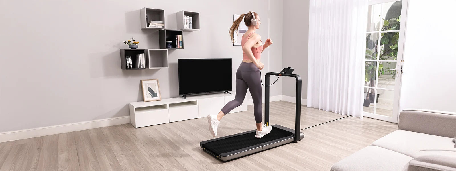 Eine Frau läuft auf einem Laufband in einem modernen Wohnzimmer mit Regalen, einem Fernseher und großen Fenstern, die natürliches Licht hereinlassen.