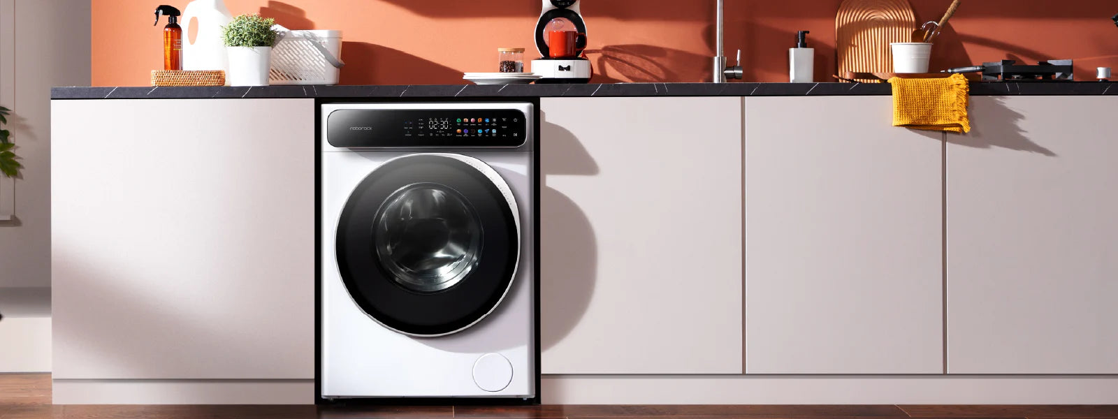 In einer Küche mit weißen Schränken und einer orangefarbenen Wand ist eine moderne Frontlader-Waschmaschine unter einer Arbeitsplatte installiert.