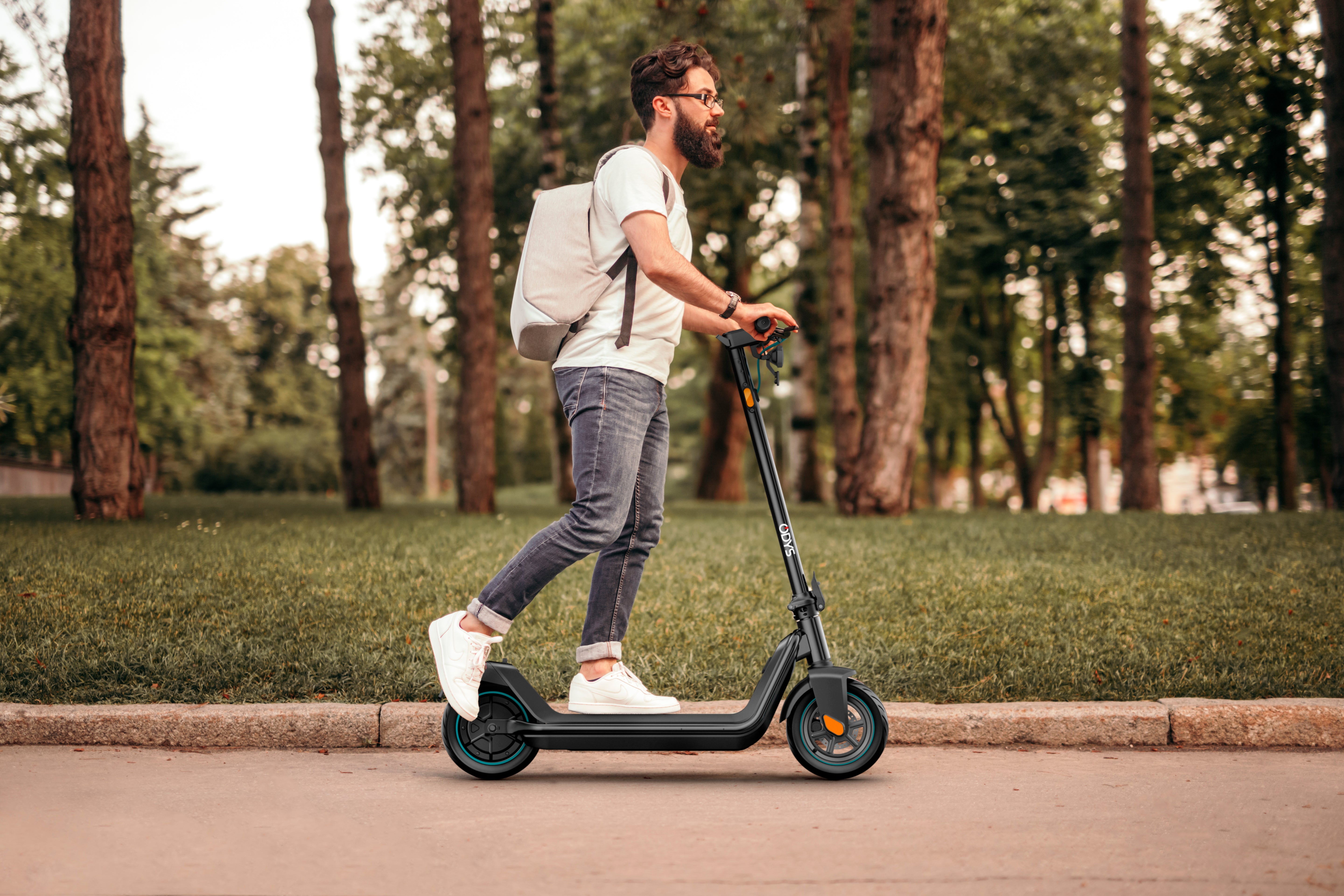 Ein Mann mit einem Rucksack fährt mit einem Odys ALPHA X5 PRO E-Scooter auf einem gepflasterten Weg neben Grasflächen und Bäumen.