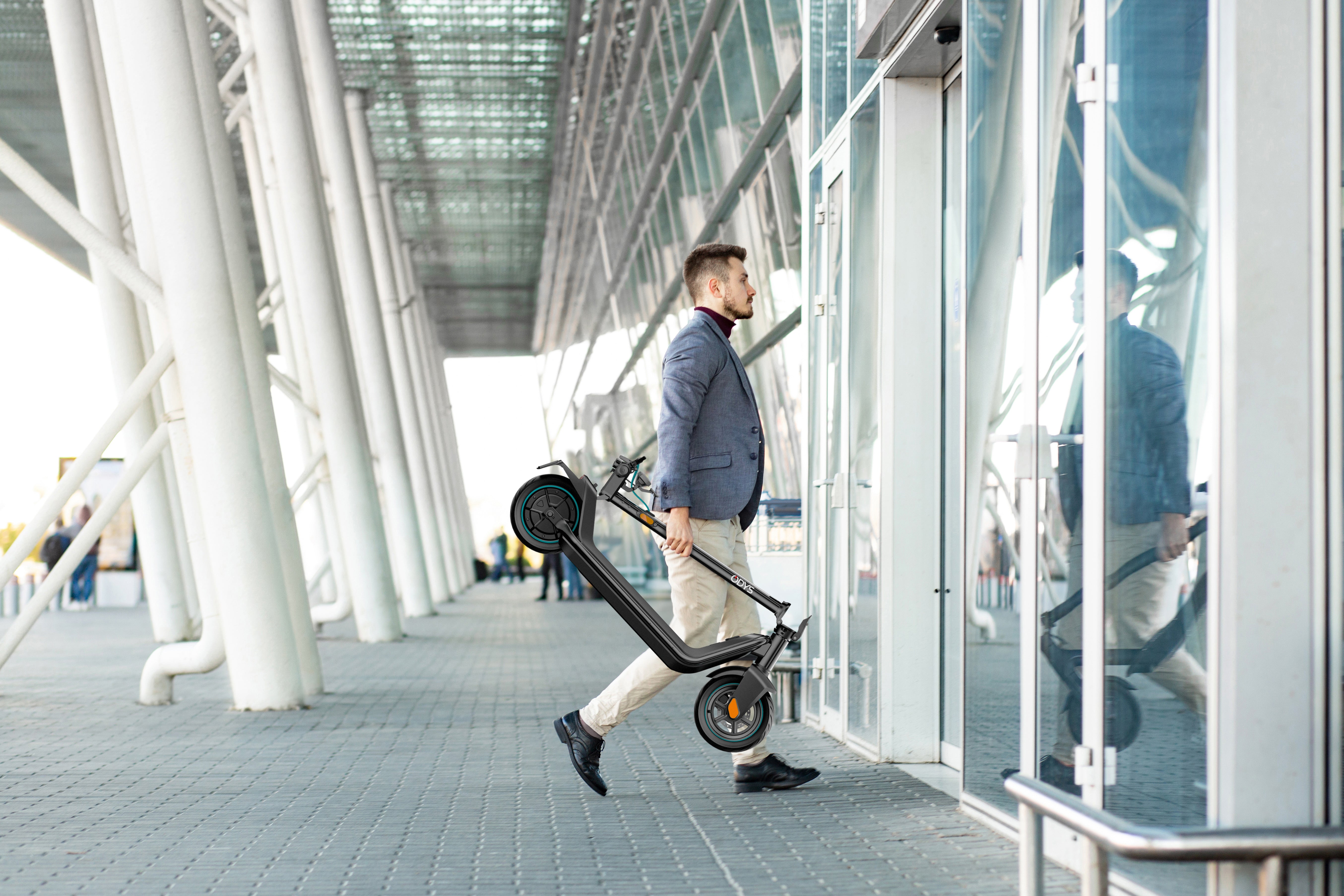 Ein Mann in Geschäftskleidung trägt einen Odys ALPHA X5 PRO E-Scooter, als er sich einer Glastür in einem modernen Gebäude nähert.