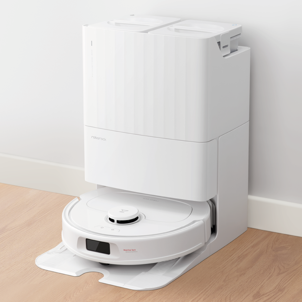 Der Roborock Qrevo Plus White Staubsaugerroboter ist in seiner selbstreinigenden Ladestation auf einem hellen Holzboden vor einer weißen Wand angedockt.