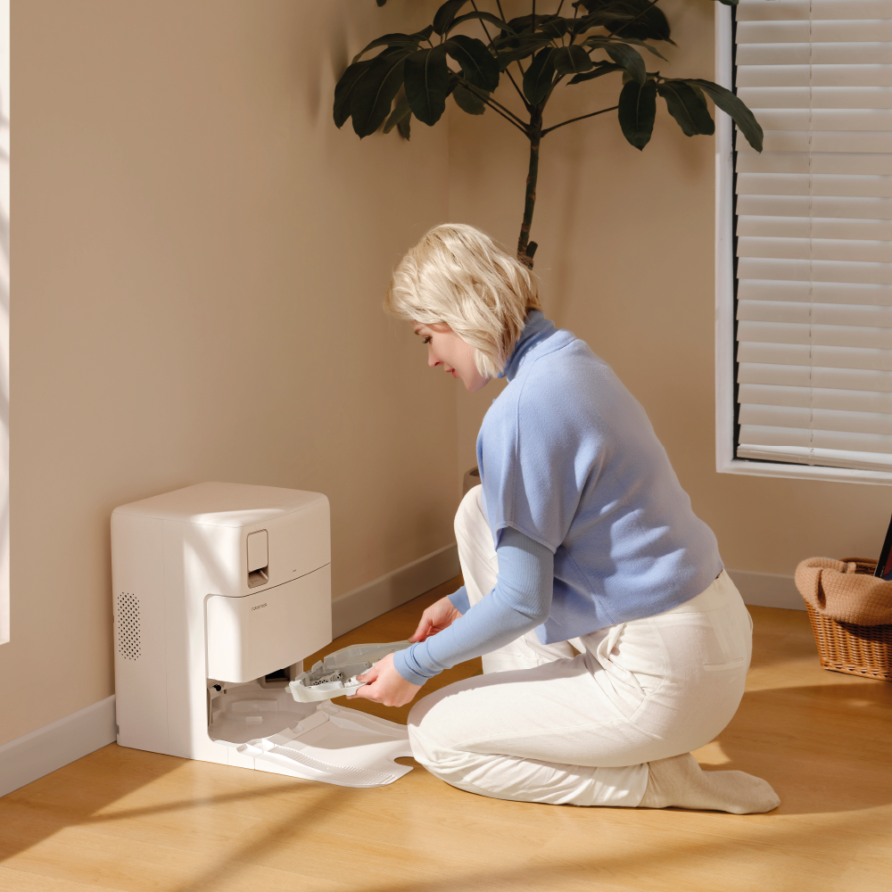 Eine leicht bekleidete Frau kniet auf einem Holzboden und nimmt ein Tablett aus dem Roborock Qrevo Plus White mit Wasseranschluss, das an der Wand neben einer Pflanze und einem Fenster steht.