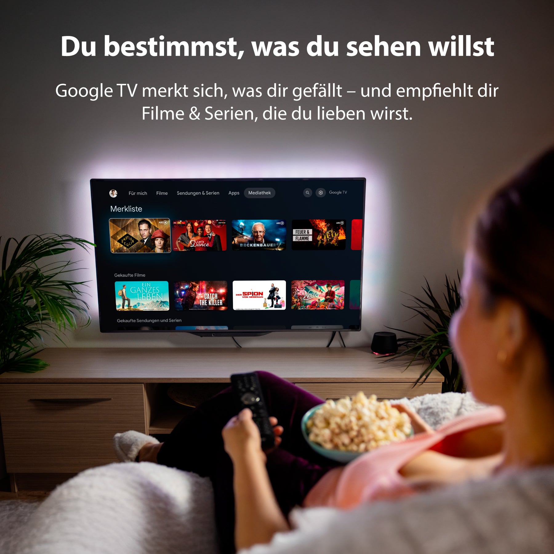 Eine Person sitzt mit Popcorn auf einer Couch, hält eine Fernbedienung in der Hand und blickt auf den Dyon nexGo 32H Smart LED TV, der die Google TV-Oberfläche mit verschiedenen Film- und Serienempfehlungen anzeigt.
