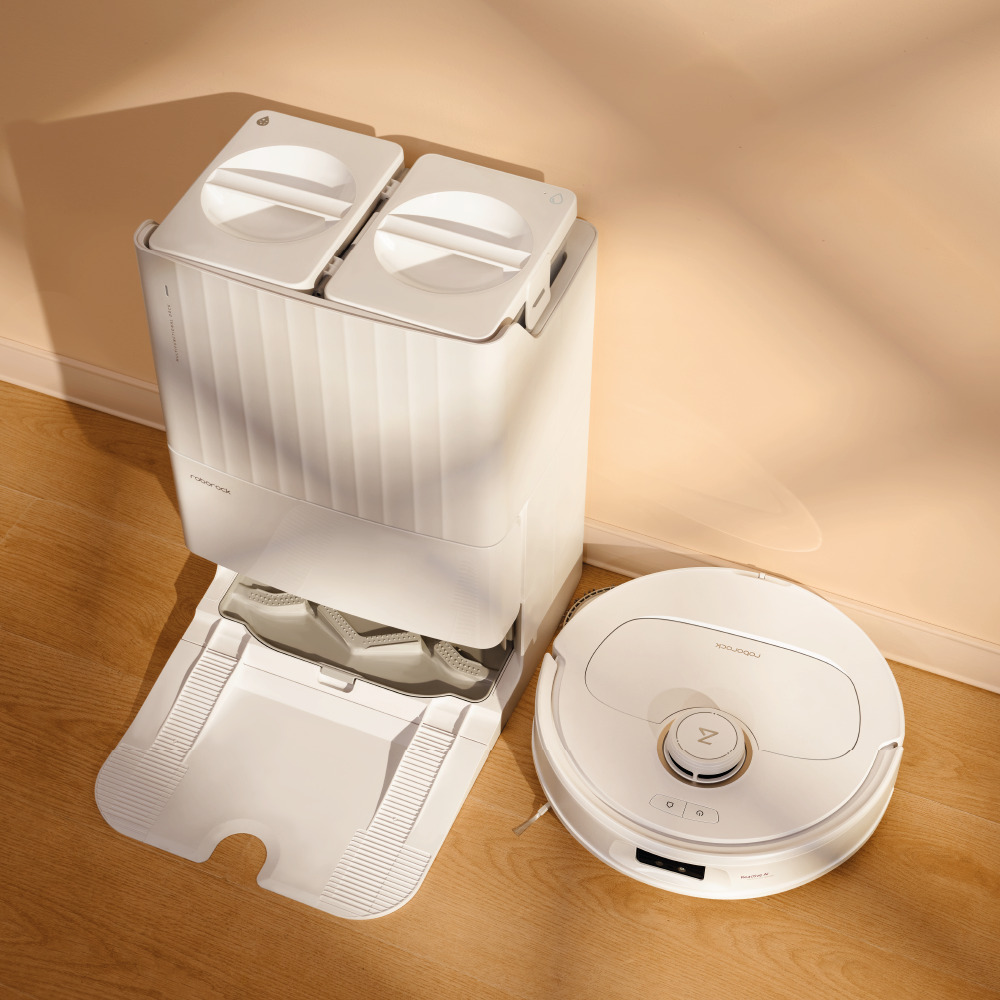 Der Roborock Qrevo MaxV White Staubsaugerroboter und seine Dockingstation mit zwei Fächern stehen auf einem hellen Holzboden vor einer beigen Wand.