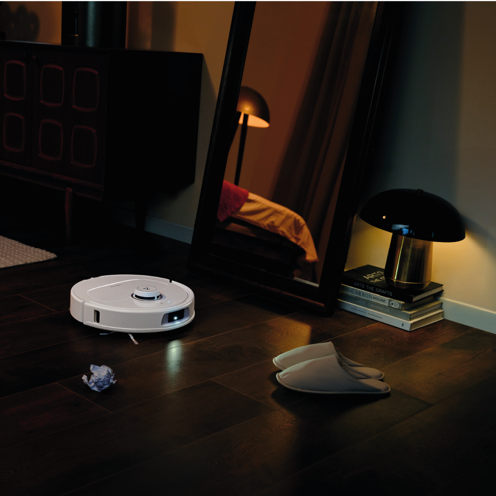 Der Roborock Qrevo MaxV White Staubsaugerroboter navigiert durch einen schwach beleuchteten Raum mit einem Spiegel, einer Lampe, gestapelten Büchern, Hausschuhen und zerknittertem Papier, die auf dem Holzboden verstreut sind.