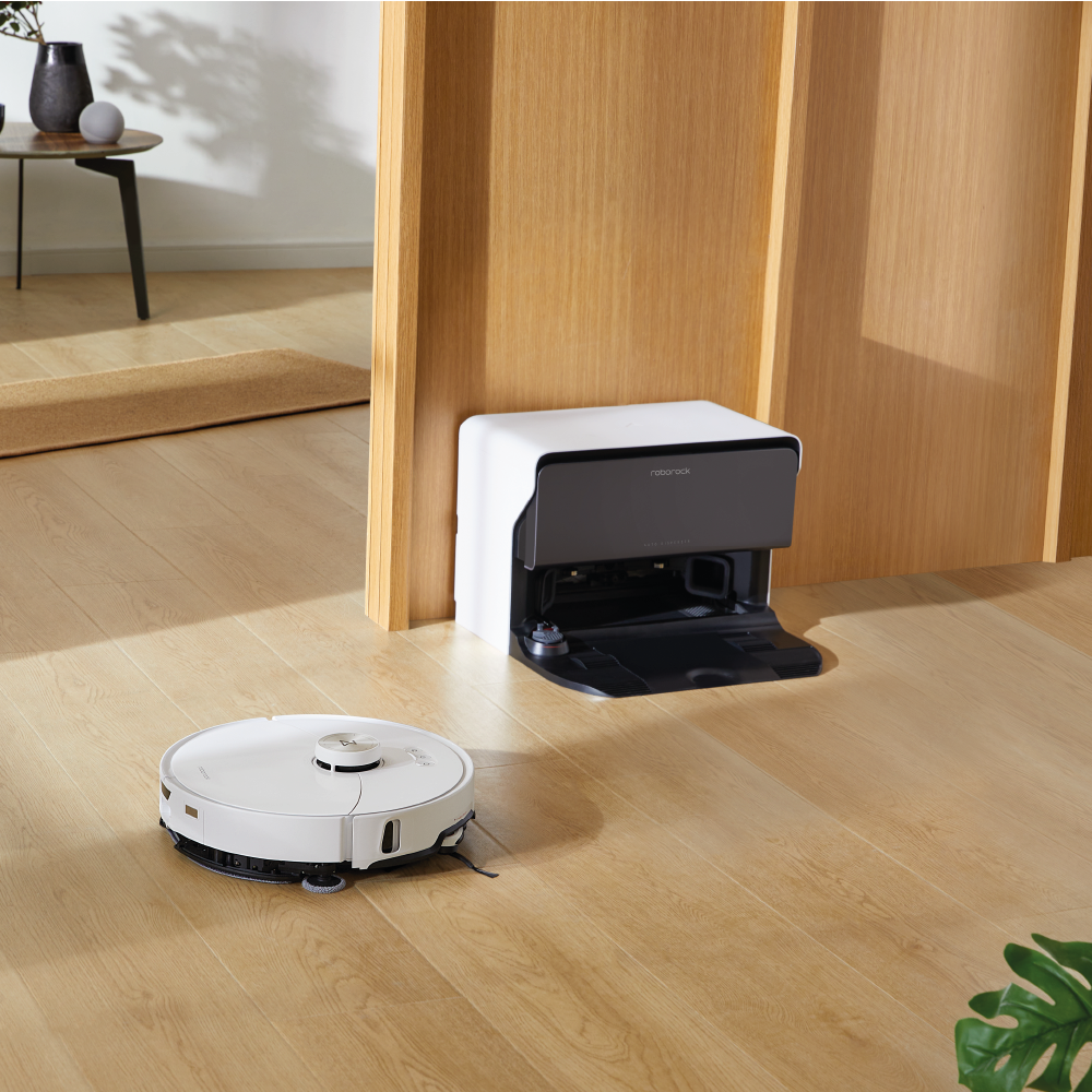Der Roborock S8 MaxV Ultra White mit Wasseranschluss bewegt sich über einen hellen Holzboden zu seiner Docking-Station, mit minimalem Mobiliar im Hintergrund.