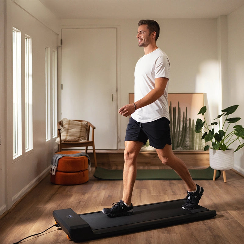 Ein Mann in Sportkleidung benutzt den Kingsmith WalkingPad A1 Pro Klappbares Laufband Schreibtisch bis 136 kg in einem hellen Wohnzimmer mit Holzboden, einem Stuhl, einer Pflanze und Kunstwerken im Hintergrund.