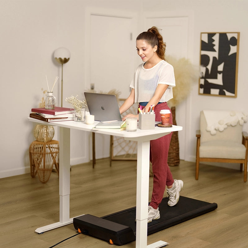 Eine Frau benutzt ein Kingsmith WalkingPad A1 Pro Klappbares Laufband Schreibtisch bis 136 kg unter einem Stehpult, während sie an ihrem Laptop in einem mit Büchern und Dekoration gefüllten Heimbüro arbeitet.