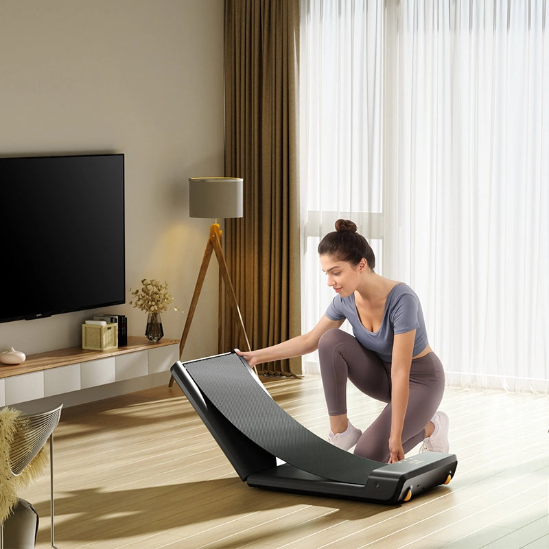 Eine Frau in Sportkleidung kniet auf einem Holzboden und entfaltet einen Kingsmith WalkingPad A1 Pro Klappbares Laufband Schreibtisch bis 136 kg in einem modernen Wohnzimmer mit Fernseher und großem Fenster.