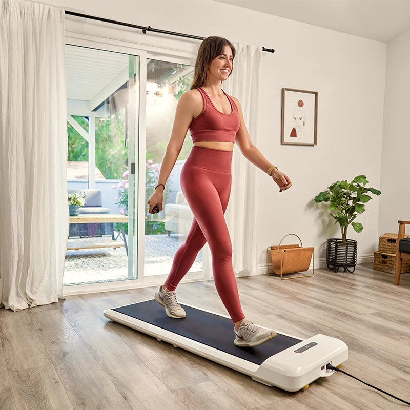 Eine Frau in passender roter Trainingskleidung benutzt ein Kingsmith WalkingPad C2 Klappbares Laufband in einem hellen Wohnzimmer mit Holzböden, einer Pflanze und Glasschiebetüren, die zu einer Terrasse führen.