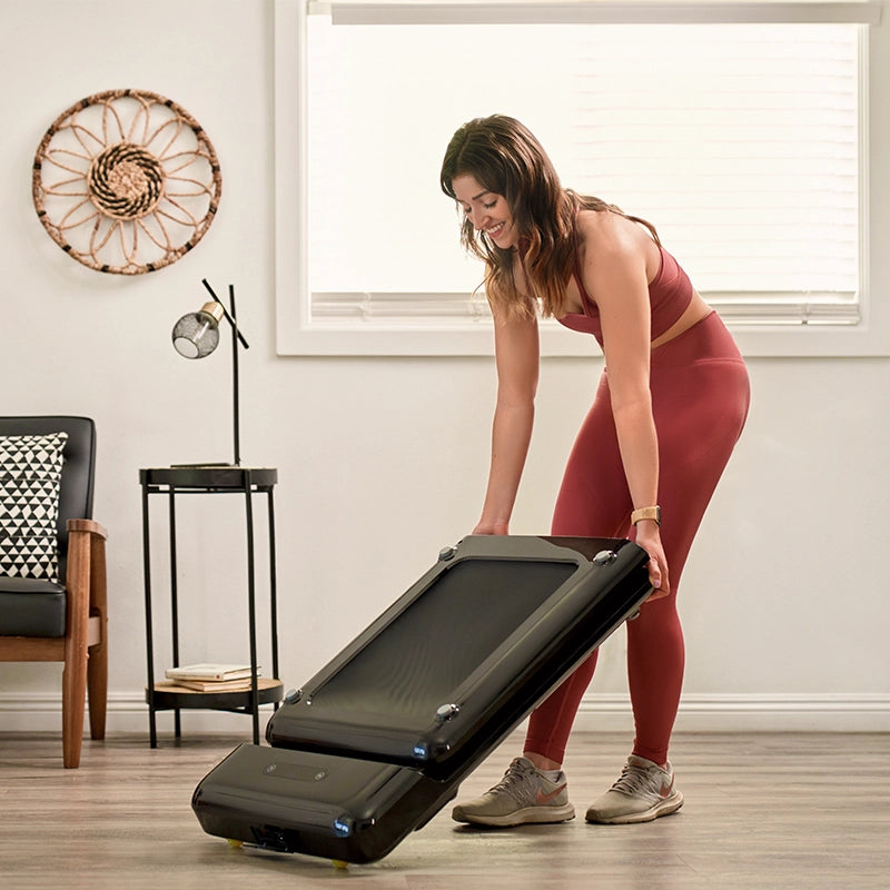 Eine Frau in Trainingskleidung faltet ein Kingsmith WalkingPad C2 Klappbares Laufband in einem hellen, modernen Wohnzimmer mit Holzboden und minimalem Dekor.