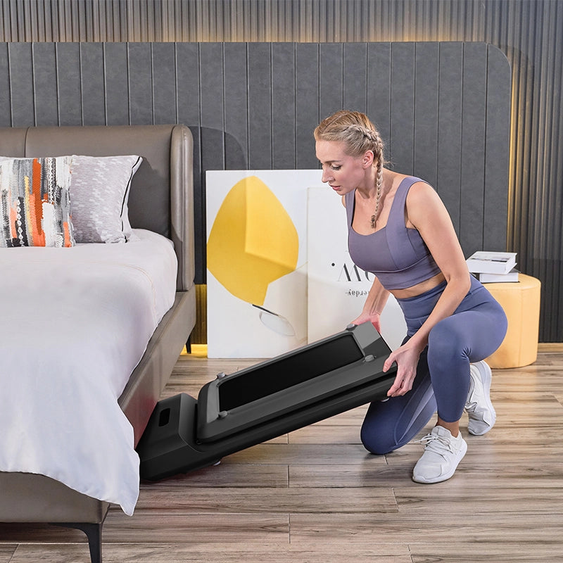 Eine Frau in Sportkleidung kniet neben einem Bett und schiebt das Kingsmith WalkingPad C2 Klappbares Laufband darunter, um es in einem modernen Schlafzimmer einfach zu verstauen.