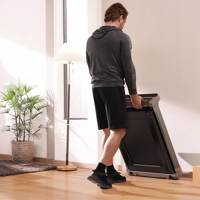Eine Person in Sportkleidung bewegt und faltet das Kingsmith WalkingPad G1 Klappbares Laufband mit Handläufen in einem hellen Raum mit Holzfußboden, einer Stehlampe und einer Topfpflanze.