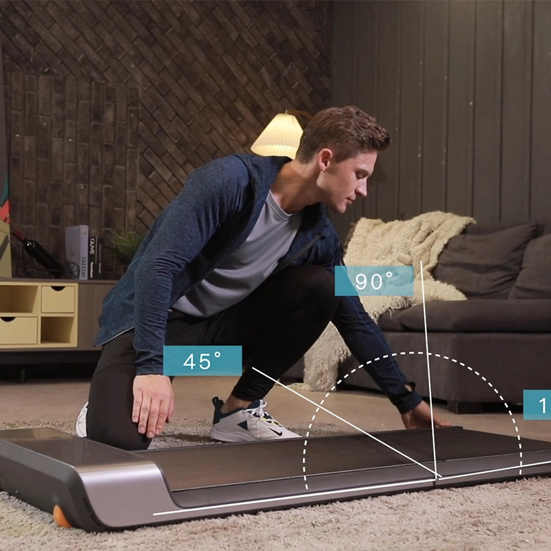 Ein Mann kniet neben einem Kingsmith WalkingPad P1 Klappbares Laufband für Schreibtisch, das auf seiner Oberfläche 45°- und 90°-Winkel misst, mit einer Couch und einer Lampe im Hintergrund.