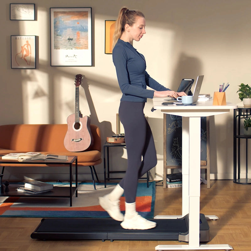 Eine Frau benutzt das Kingsmith WalkingPad Z1 Klappbares Laufband für Schreibtisch unter ihrem Stehpult, während sie an ihrem Laptop in einem mit einer Gitarre und Kunstwerken dekorierten Wohnzimmer arbeitet.