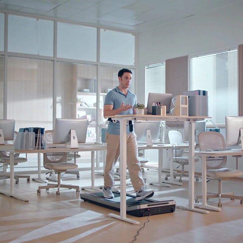 Ein Mann arbeitet an einem Stehpult und läuft dabei auf einem Kingsmith WalkingPad Z3 Hybrid+ Klappbares Laufband für Schreibtisch in einem modernen Großraumbüro mit mehreren leeren Schreibtischen und Computern.