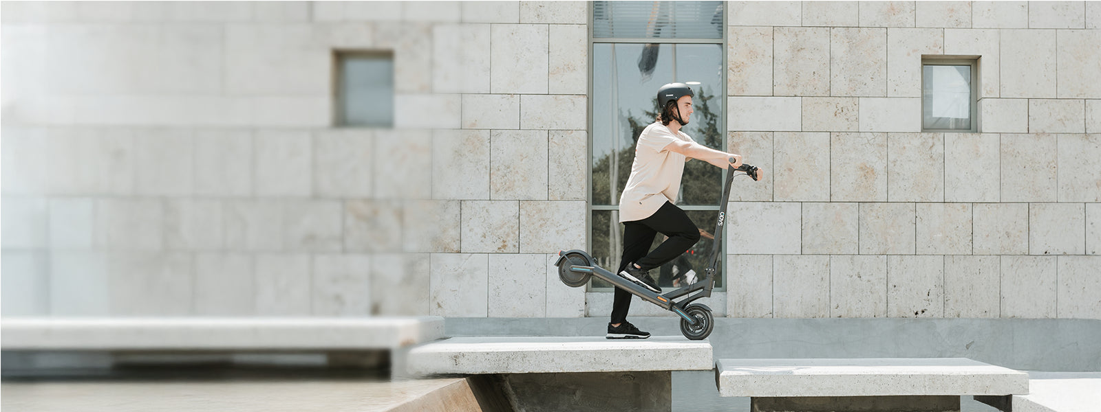 Eine Person, die einen Helm trägt, fährt mit einem Elektroroller auf einer Betonfläche vor einem modernen Steingebäude mit Fenstern.