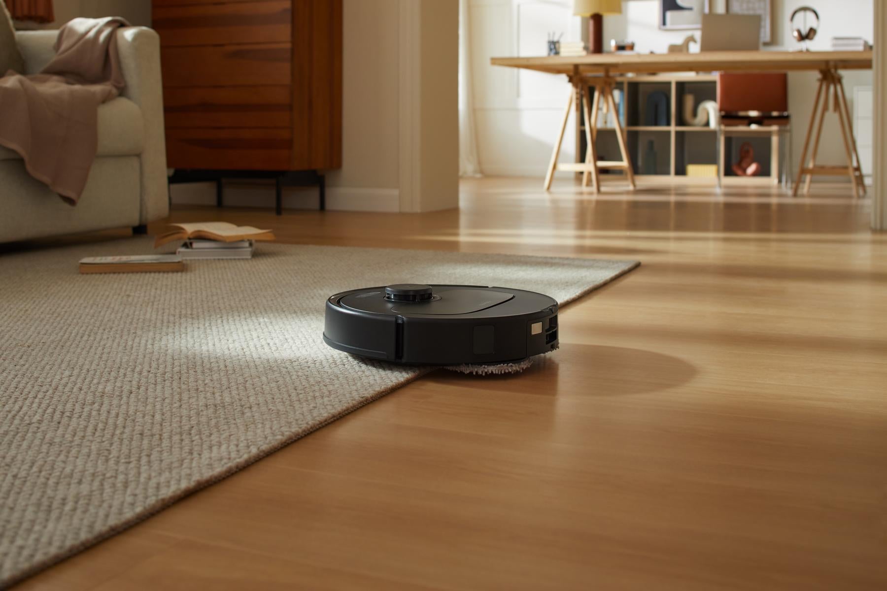 Ein schwarzer Roborock Qrevo 598 Staubsauger mit HyperForce-Saugfunktion wechselt in einem Wohnzimmer mit einem Sofa, einem Bücherregal und einem Schreibtisch im Hintergrund von Teppich auf Holzboden.