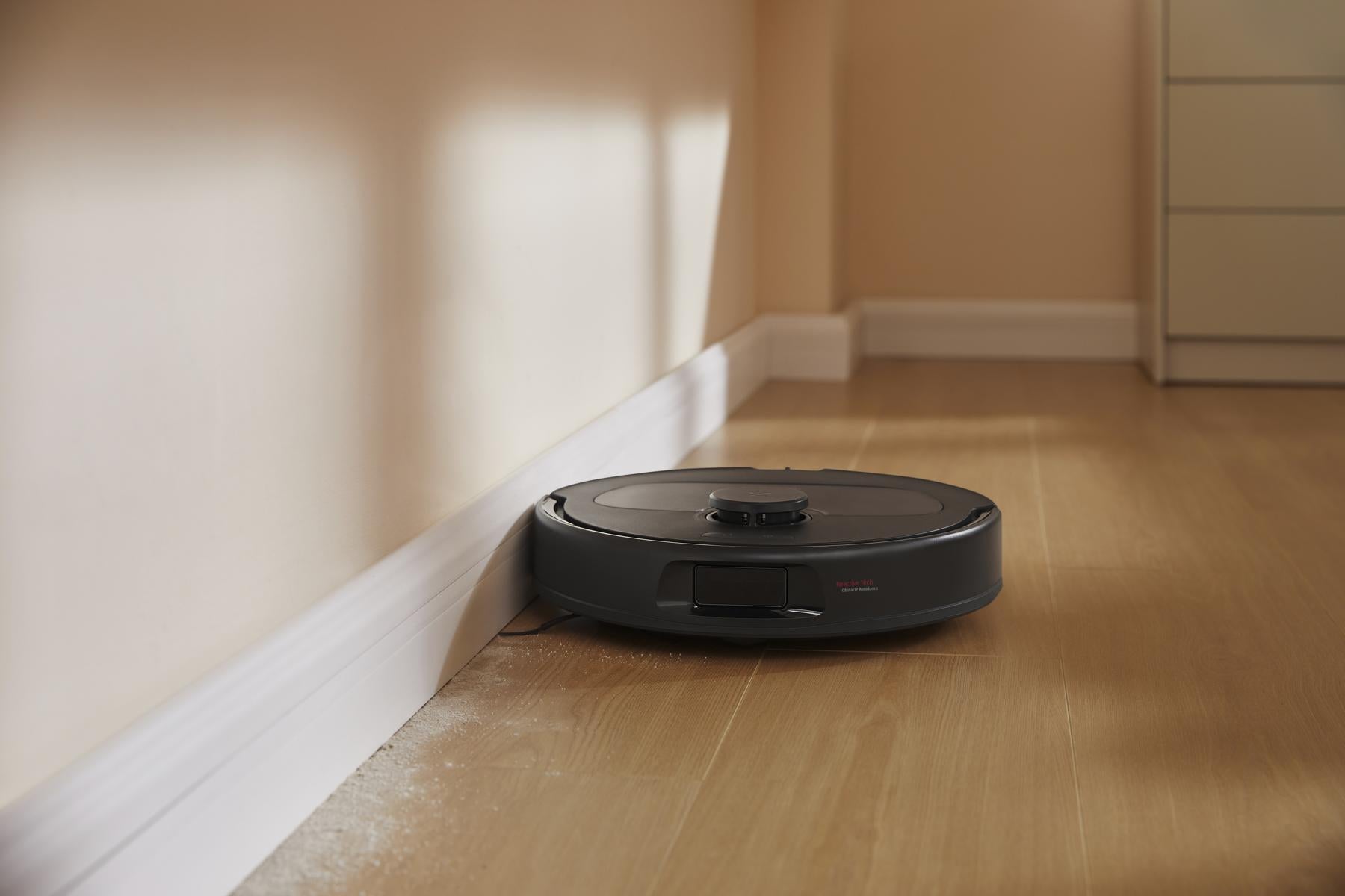 Der Roborock Qrevo 598 Black Saugroboter mit HyperForce-Saugfunktion gleitet über einen hellen Holzfußboden in der Nähe einer weißen Wand und reinigt effizient Schmutz.