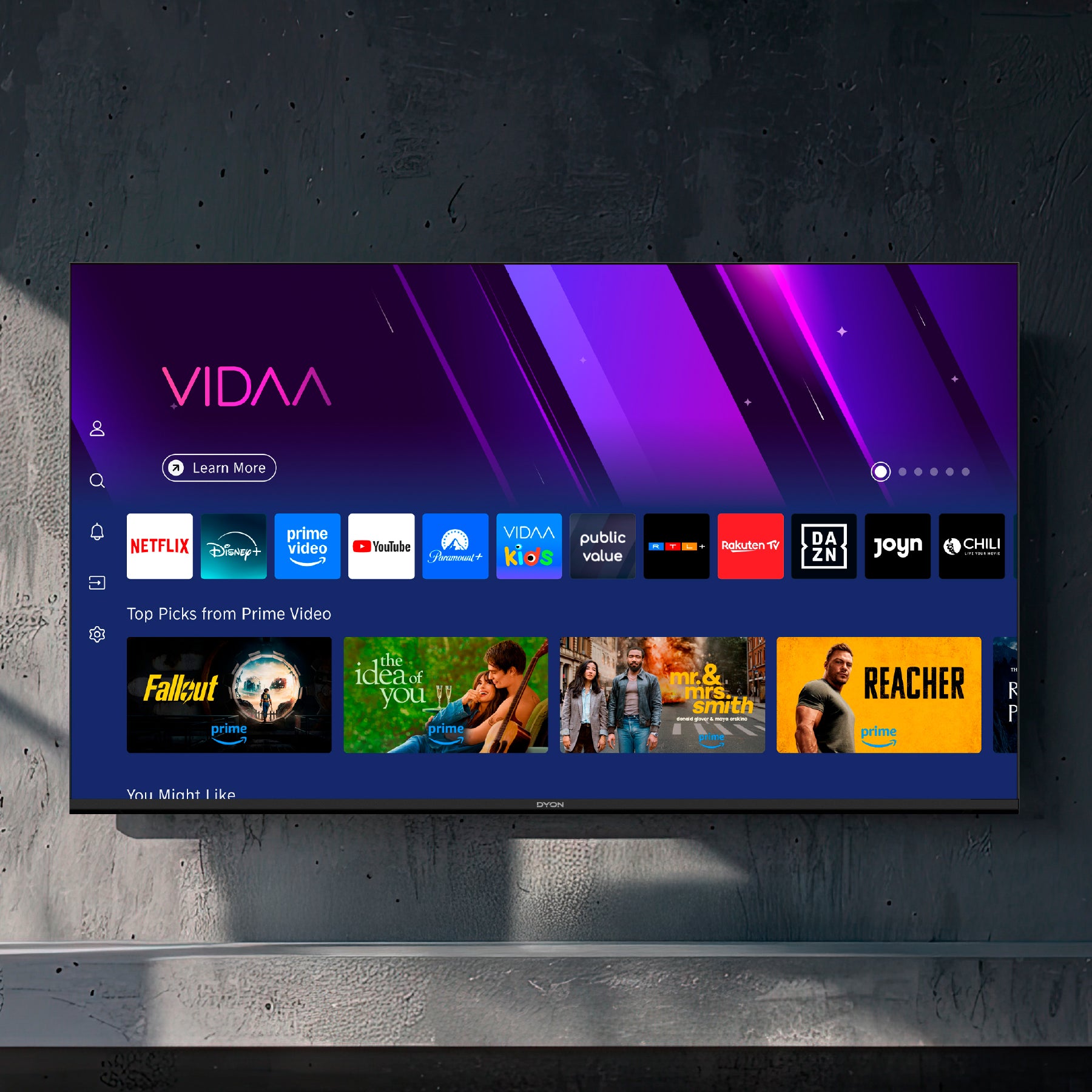 Ein Dyon Smart 43 VX Smart LED-Fernseher wird an der Wand montiert und zeigt die VIDAA Smart TV-Oberfläche mit mehreren Streaming-Apps und Show-Empfehlungen an.
