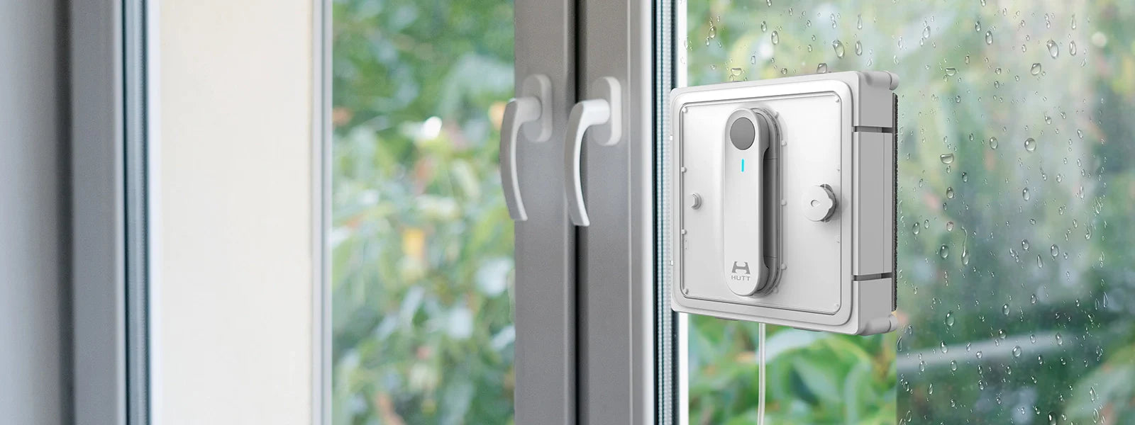 Ein Fensterreinigungsroboter ist an einem Glasfenster angebracht und reinigt die Oberfläche, während auf der Außenseite Regentropfen zu sehen sind. In der Nähe sind zwei weiße Fenstergriffe zu sehen.