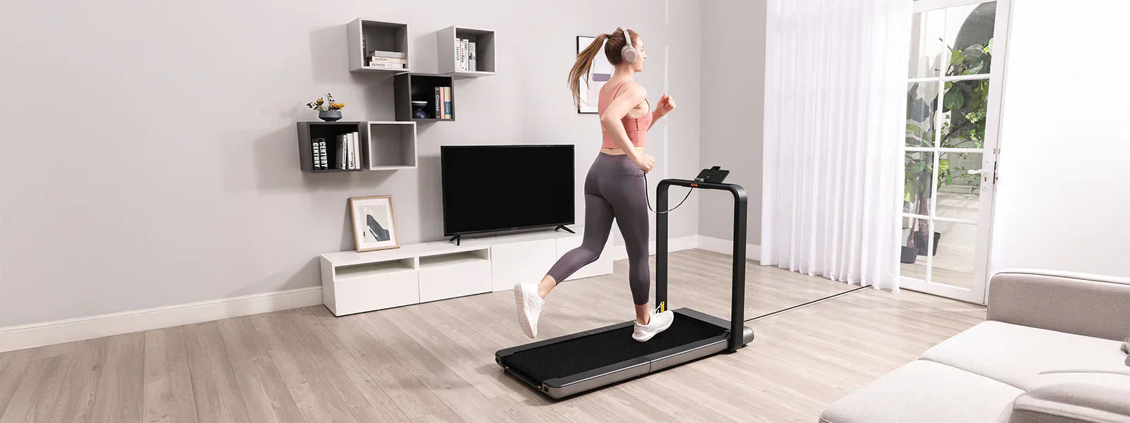 Eine Frau läuft auf einem Laufband in einem modernen Wohnzimmer, sie trägt Sportkleidung und Kopfhörer. Im Hintergrund sind ein Fernseher, Regale und ein Fenster mit durchsichtigen Vorhängen zu sehen.