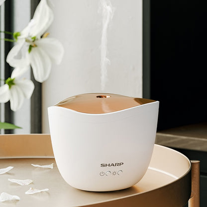 Ein Sharp Ultraschall Aroma Zerstaeuber von Sharp verströmt Nebel auf einem Tablett, umgeben von Blütenblättern und einer weißen Lilie, und erinnert mit seiner eleganten Duftverbreitung an Aromatherapie.