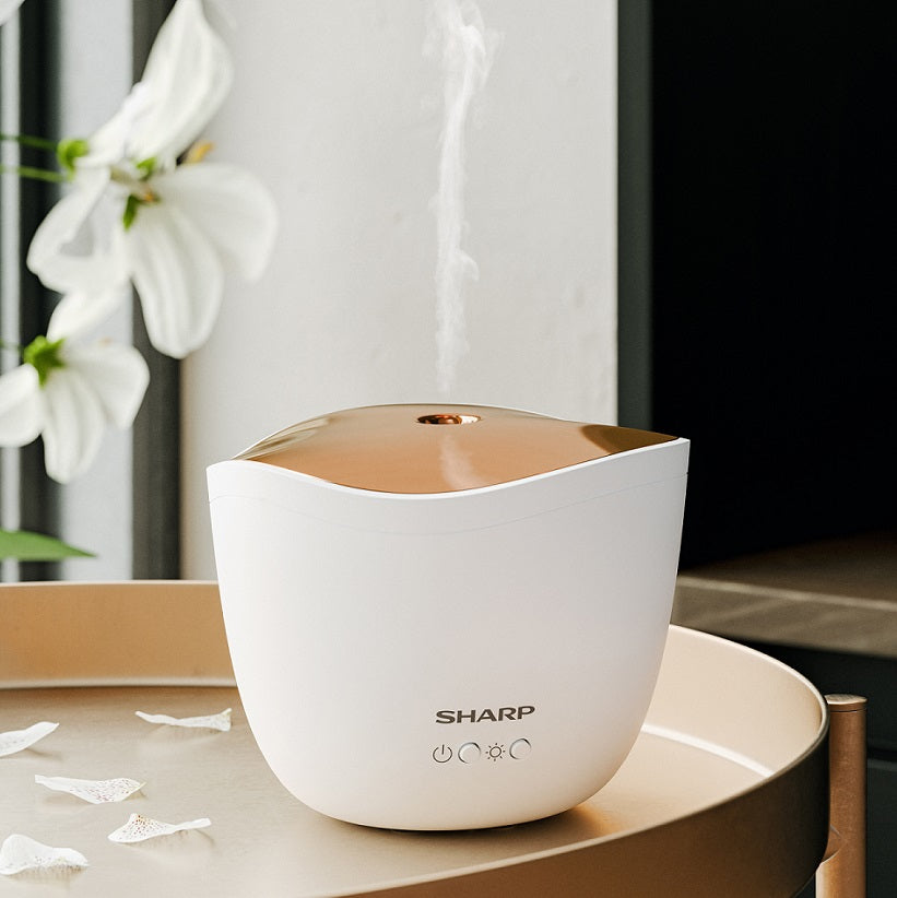 Ein Sharp Ultraschall Aroma Zerstaeuber von Sharp verströmt Nebel auf einem Tablett, umgeben von Blütenblättern und einer weißen Lilie, und erinnert mit seiner eleganten Duftverbreitung an Aromatherapie.