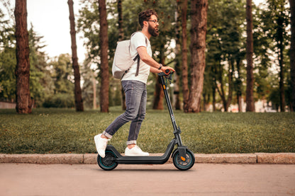 Ein Mann mit einem Rucksack fährt mit einem Odys ALPHA X5 PRO E-Scooter auf einem gepflasterten Weg neben Grasflächen und Bäumen.