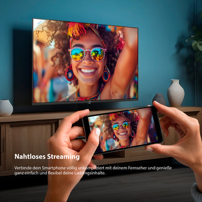 Eine Person benutzt ein Smartphone, um eine lächelnde Frau auf einen Dyon Ultimax 40F-TI Fernseher zu spiegeln; beide Bildschirme zeigen das gleiche lebendige Bild. Der deutsche Text hebt das nahtlose Streaming mit dem Dyon Ultimax 40F-TI hervor.