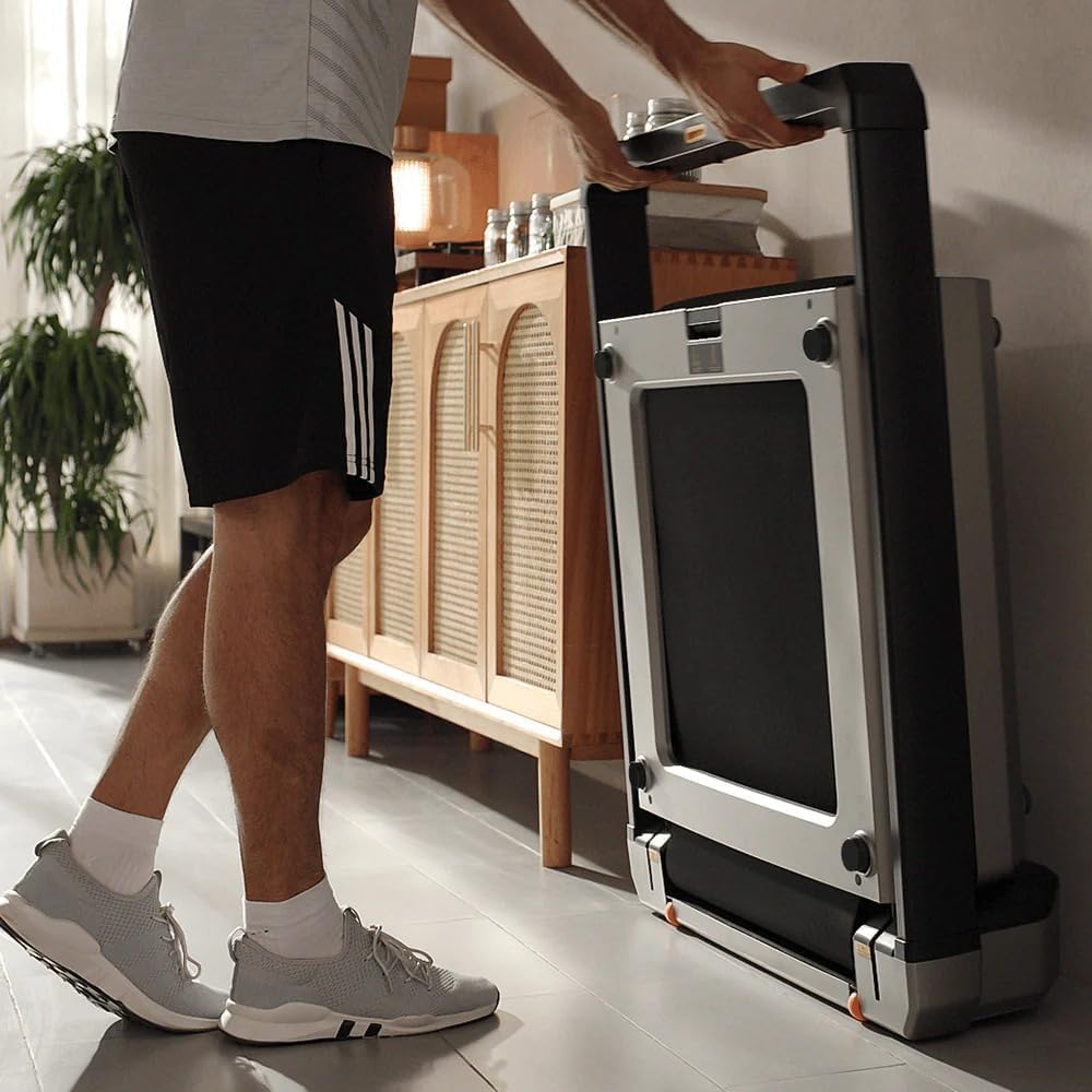 Eine Person in Sportkleidung steht neben einem Kingsmith WalkingPad X23/MX16 Klappbares Laufband (bis 16 km/h & 136 kg), das aufrecht an einer Wand in einer gemütlichen Wohnumgebung steht.