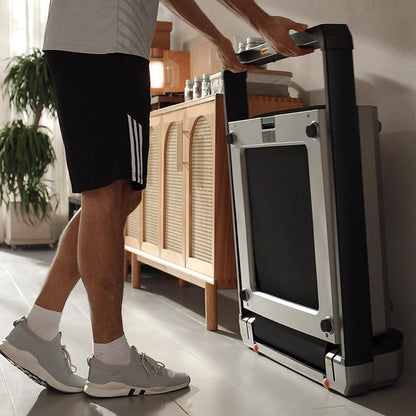 Eine Person in Sportkleidung steht neben einem Kingsmith WalkingPad X23/MX16 Klappbares Laufband (bis 16 km/h & 136 kg), das aufrecht an einer Wand in einer gemütlichen Wohnumgebung steht.
