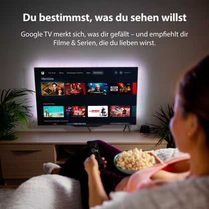 Eine Person sitzt mit Popcorn auf einer Couch, hält eine Fernbedienung in der Hand und blickt auf den Dyon nexGo 32H Smart LED TV, der die Google TV-Oberfläche mit verschiedenen Film- und Serienempfehlungen anzeigt.