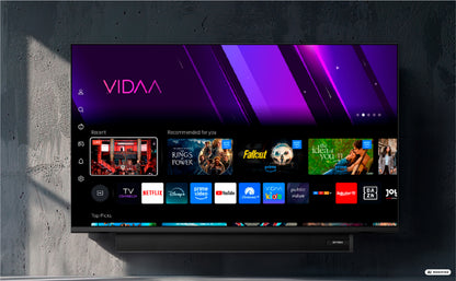 Ein DYON Movie Smart 32 VX-S 32-Zoll-HD-Smart-TV – Soundboost von Dyon ist an einer grauen Wand montiert und zeigt die VIDAA-Smart-TV-Benutzeroberfläche mit verschiedenen Streaming-Apps und empfohlenen Inhalten an.
