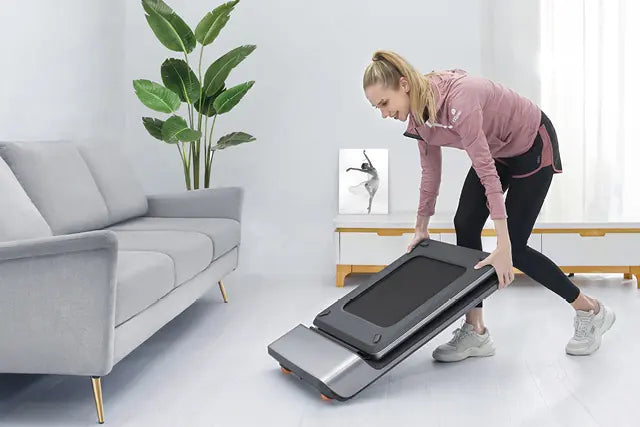 Eine Frau in Sportkleidung entfaltet ein kompaktes Laufband in einem modernen Wohnzimmer mit einem Sofa, einer Topfpflanze und einem gerahmten Kunstwerk.