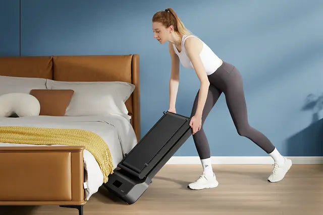 Eine Frau in Sportkleidung faltet und hebt ein kompaktes Laufband neben einem Bett in einem modernen Schlafzimmer.