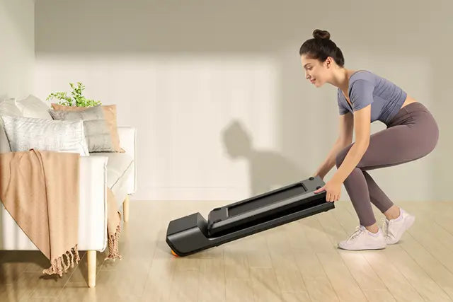 Eine Frau in Sportkleidung hebt ein zusammengeklapptes kompaktes Laufband vom Boden in einem hellen Wohnzimmer mit einem Sofa und Kissen im Hintergrund.