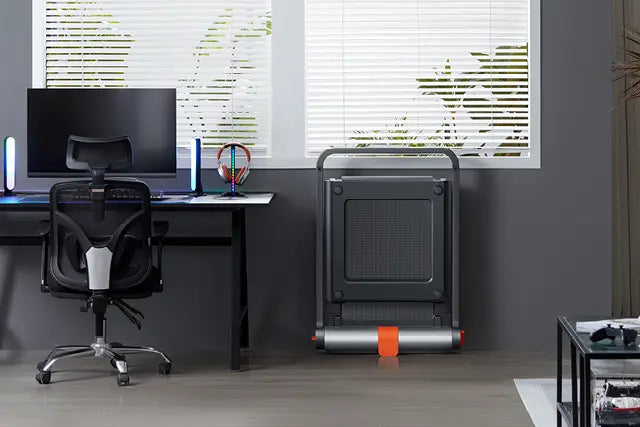 Ein modern eingerichteter Schreibtisch mit einem Monitor, einem Bürostuhl und einem zusammengefalteten Laufband, das an der Wand in der Nähe eines Fensters mit Jalousien steht.