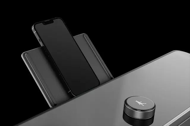 Ein schwarzes Smartphone ruht aufrecht in einer kabellosen Ladestation neben einem runden Bedienknopf auf einer glatten, reflektierenden Oberfläche.