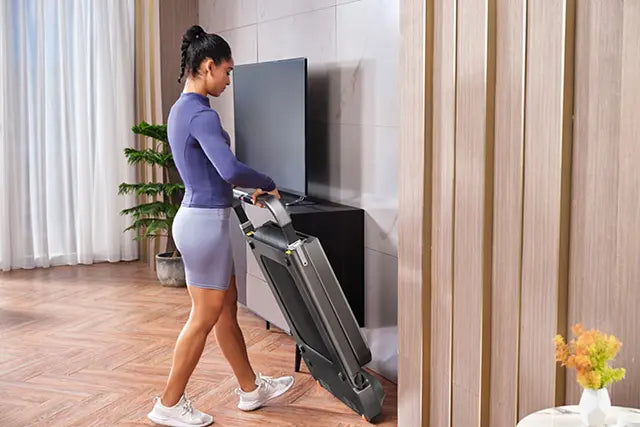 Eine Frau in Sportkleidung lagert ein zusammengeklapptes Laufband aufrecht an einer Wand in einem modernen, gut beleuchteten Wohnzimmer.