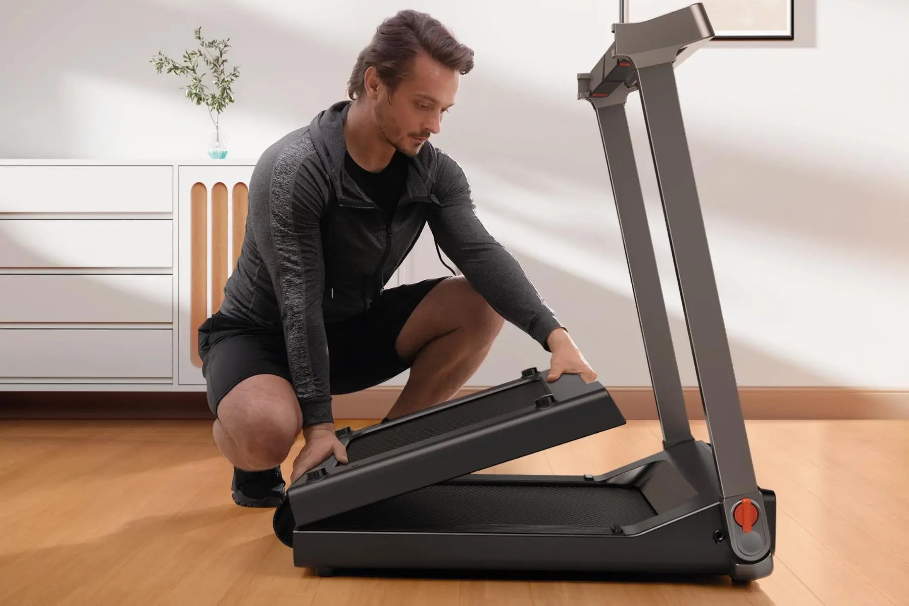 Ein Mann in Sportkleidung kniet auf einem Holzboden und klappt ein kompaktes Laufband in einem modernen, gut beleuchteten Raum aus.