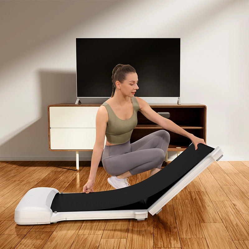 Eine Frau in Trainingskleidung entfaltet das Kingsmith WalkingPad C1 Klappbares Laufband in ihrem Wohnzimmer mit Holzboden und Fernseher - ideal für Homeoffice Fitness.