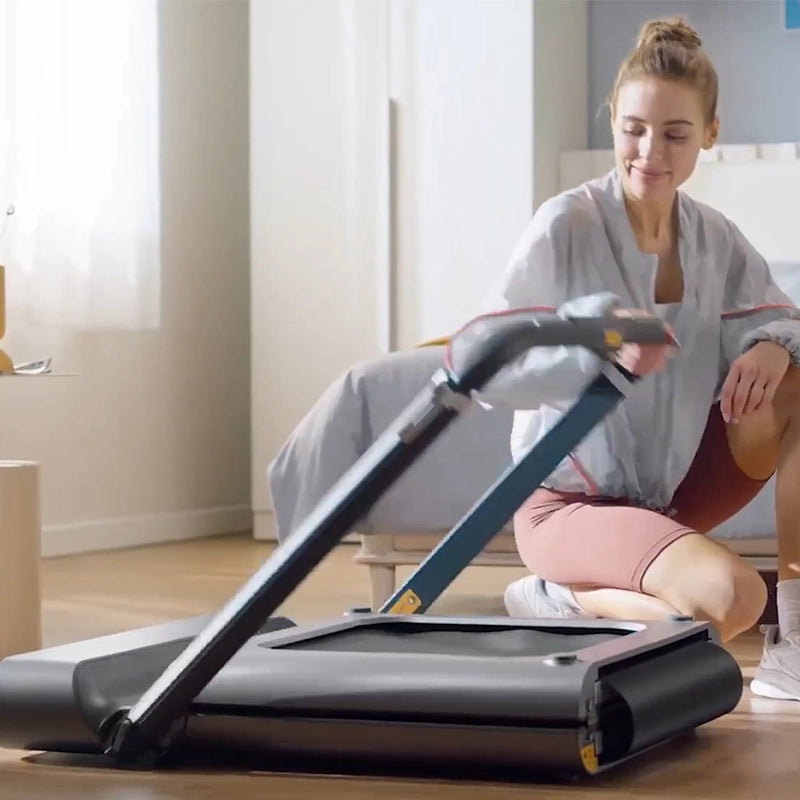Eine Frau kniet auf dem Boden und klappt das Kingsmith WalkingPad R1H/R1 Pro 2IN1 klappbares Laufband in einem hellen, modernen Schlafzimmer zusammen.