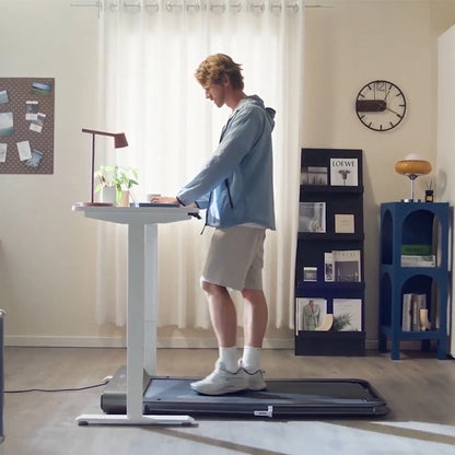 Ein Mann benutzt einen Laptop an einem Stehpult, während er auf dem Kingsmith WalkingPad R1H/R1 Pro 2IN1 Klappbares Laufband in einem Heimbüro mit Regalen, einer Uhr und natürlichem Licht aus dem Fenster läuft.