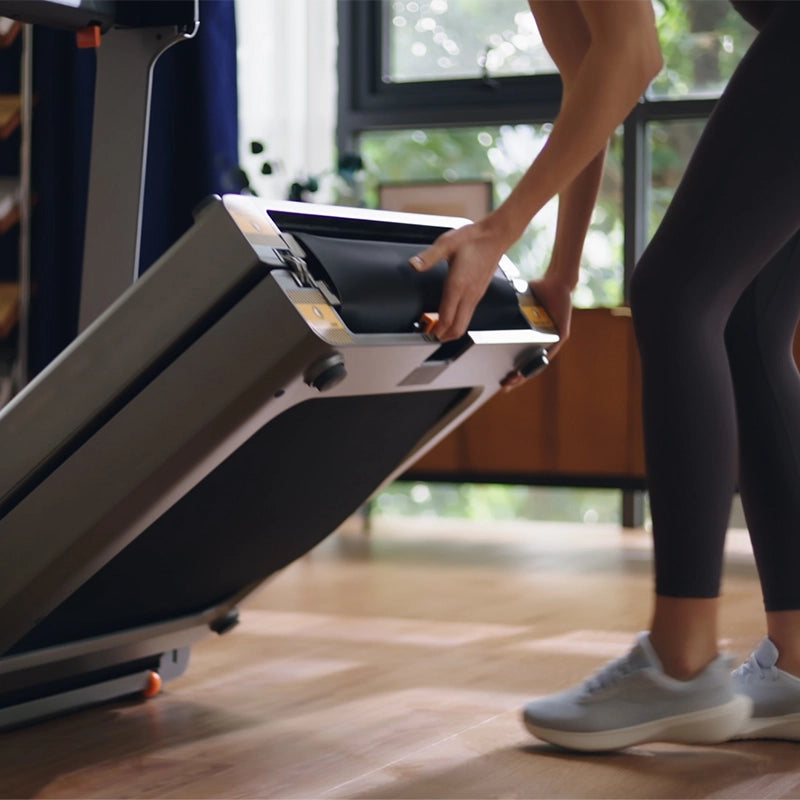 Eine Person in Sportkleidung klappt das Kingsmith WalkingPad X218 Klappbares Laufband in einem hellen, modernen Wohnzimmer mit Holzboden und Zimmerpflanzen zusammen.