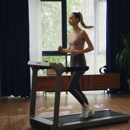 In einem hellen, modernen Wohnzimmer mit Holzfußboden und großen Fenstern läuft eine Frau in Sportkleidung auf dem Kingsmith WalkingPad X218 Klappbares Laufband und macht intensives Training zu ihrer täglichen Routine.