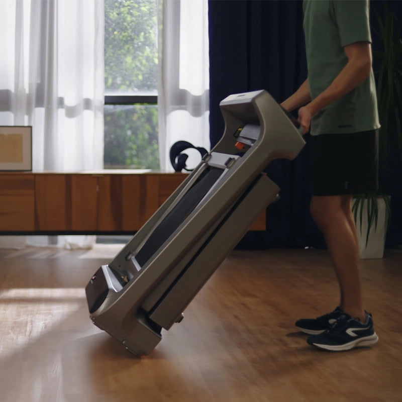 Eine Person kippt und rollt das Kingsmith WalkingPad X218 Klappbares Laufband, ein elegantes Laufband für zu Hause, über einen Holzboden in einem sonnenbeschienenen Raum.