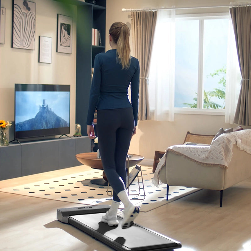 Eine Frau benutzt das Kingsmith WalkingPad Z1 Klappbares Laufband für Schreibtisch in ihrem Wohnzimmer. Sie trainiert, während sie in einer gemütlichen Umgebung mit einem Sofa, einem Couchtisch, einem großen Fenster und gerahmter Kunst fernsieht.